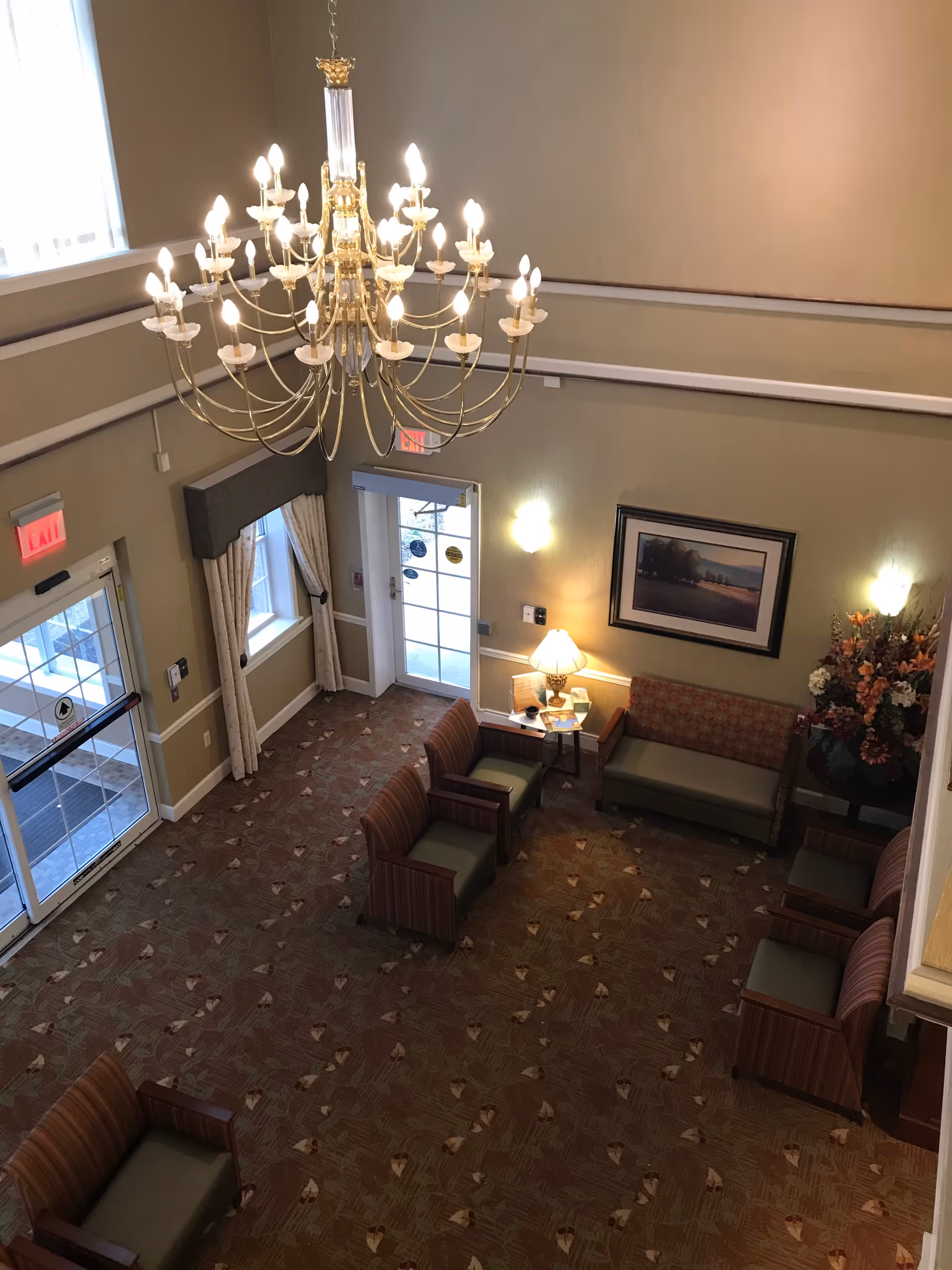 View down into a senior living facility lobby showing a large chandelier, seating area with chairs and couches, entrance doors, and a table lamp.