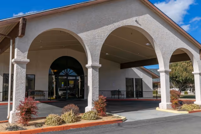 Entrance of a building with white stucco walls and large arches supported by columns. There are small bushes and plants in front of the entrance, and a covered driveway area. The sky is clear and blue.