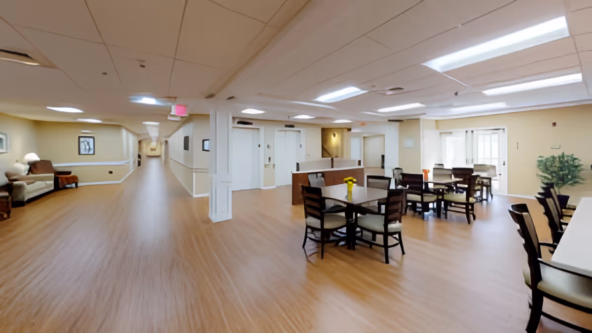 A spacious interior area of a senior living facility featuring a long hallway with seating along the left side and a dining area with tables and chairs on the right. The space has wood flooring, beige walls, and ceiling lights. There are elevators and a reception desk in the background.