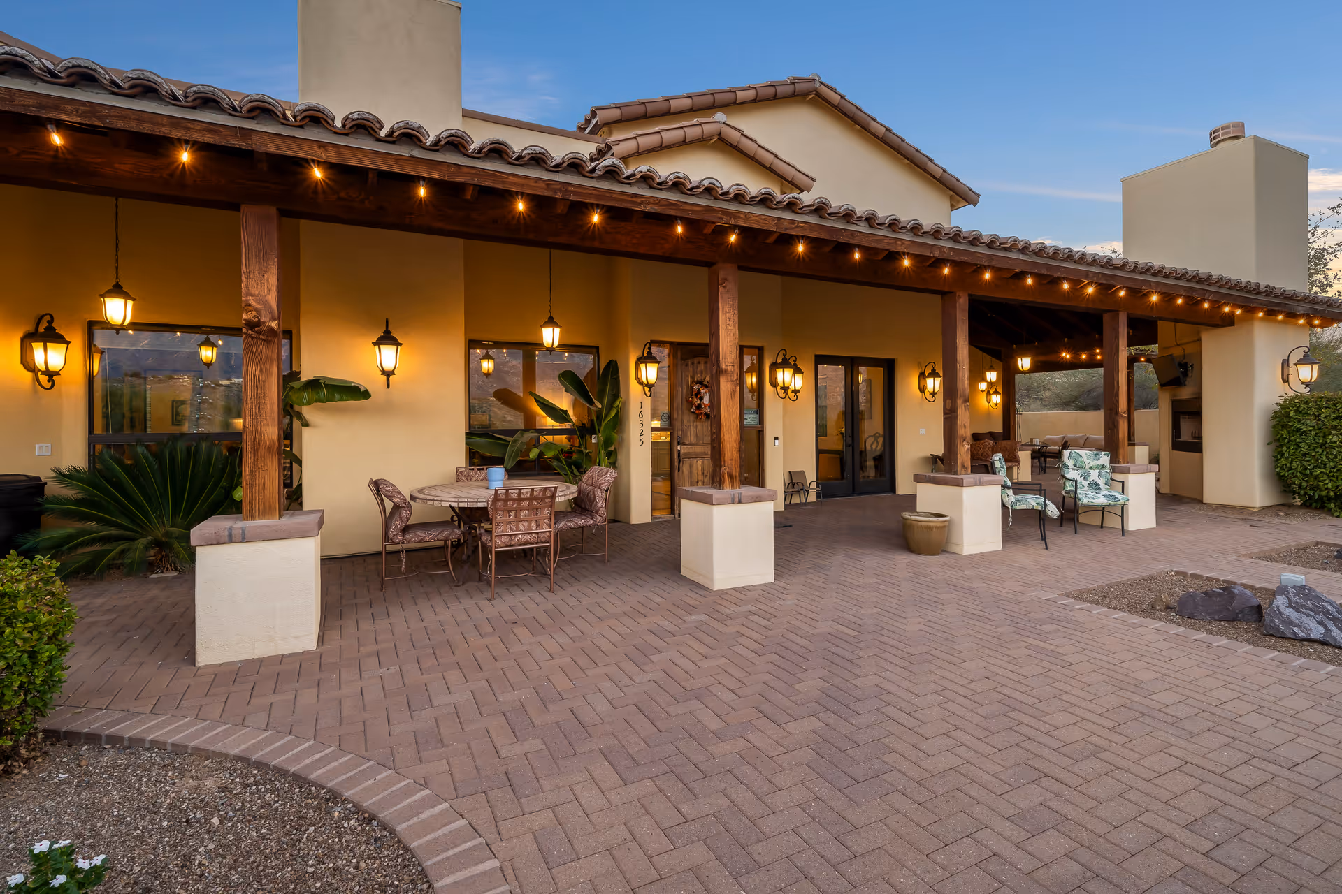 Outdoor patio area of Heritage Manor Assisted Living featuring a covered seating area with wooden beams and hanging lantern-style lights. There are several chairs and tables arranged for relaxation and socializing, with potted plants and decorative greenery around. The building has a stucco exterior with a tiled roof and a chimney, and the patio is paved with brick-like tiles.