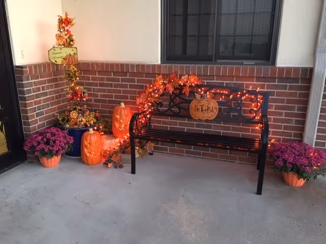 A black metal bench decorated with orange autumn leaves and orange string lights, with a wooden pumpkin sign that says 'Be Thankful'. To the left of the bench are two carved pumpkins with lights inside, a tall plant with fall decorations, and a pot of purple flowers. Another pot of purple flowers is on the right side of the bench. The setting is a covered outdoor porch area with brick walls and a concrete floor.