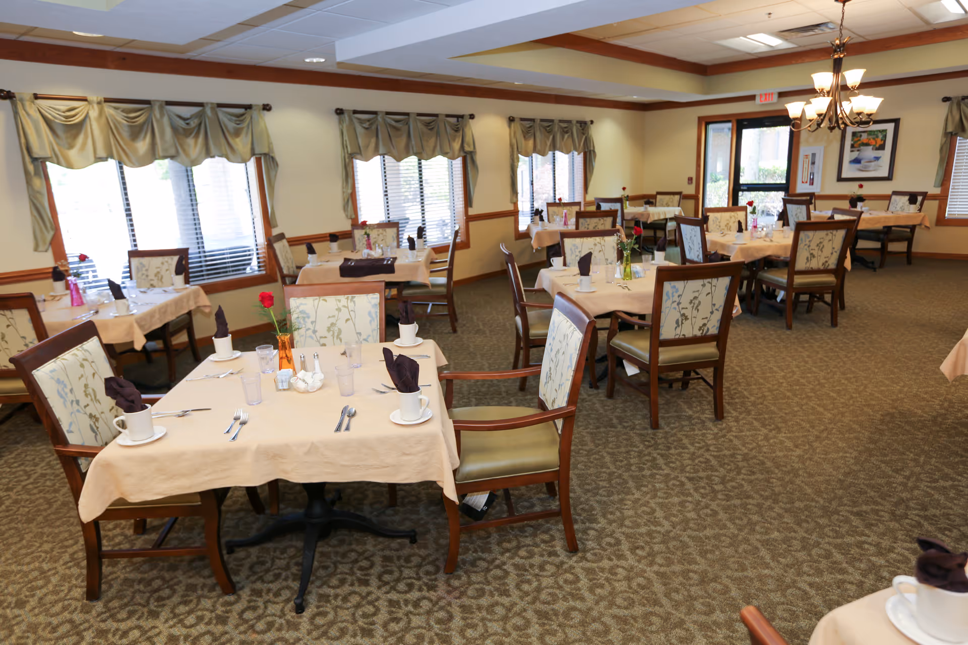 A dining room with multiple tables covered with beige tablecloths, each set with cups, glasses, silverware, and napkins folded inside cups. The chairs have wooden frames with floral patterned upholstery. The room has large windows with beige curtains, carpeted floors, and a chandelier hanging from the ceiling.