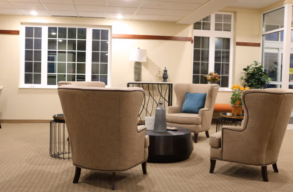 Cozy senior living lounge with upholstered armchairs arranged around a central coffee table and decorative plants and lamps.