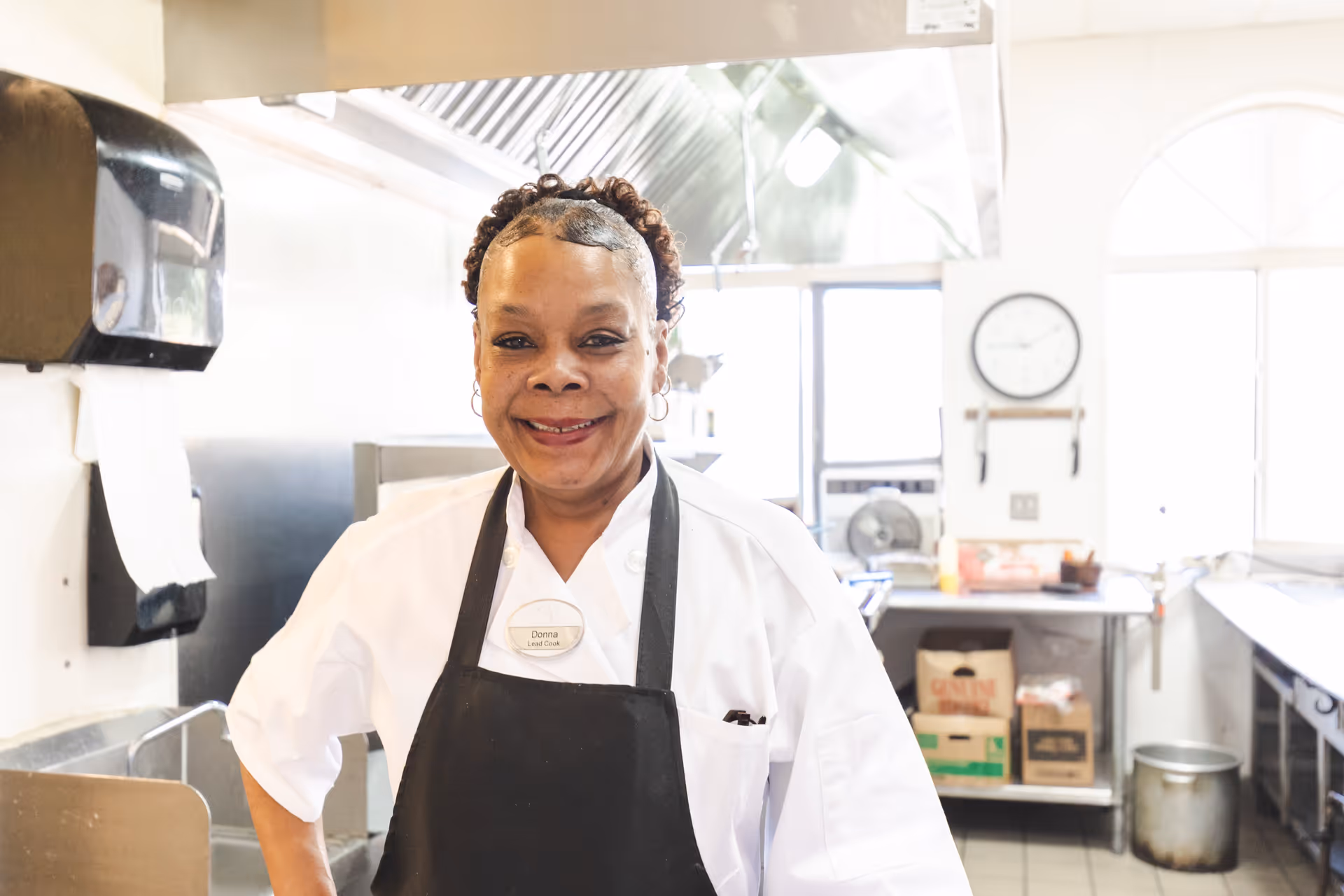A smiling woman wearing a white chef's coat and black apron stands in a commercial kitchen. She has a name tag that reads 'Donna Lead Cook'. The kitchen has stainless steel appliances, a paper towel dispenser, a clock on the wall, and various kitchen supplies in the background.