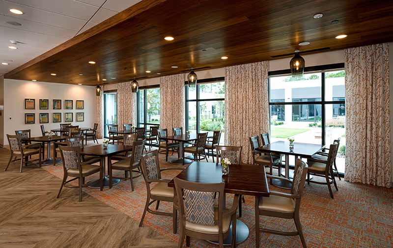Dining area with several wooden tables and upholstered chairs, pendant lights, large windows with patterned curtains, and a view of a courtyard.
