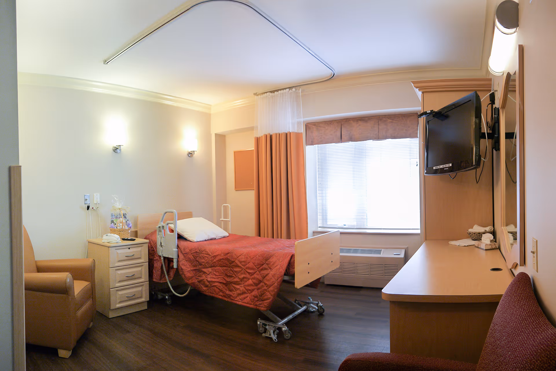 A single patient room in a care center featuring a hospital bed with a red quilt, a bedside table with a gift basket and telephone, a comfortable armchair, a wall-mounted TV, a desk with a mirror, and a window with blinds and curtains letting in natural light.