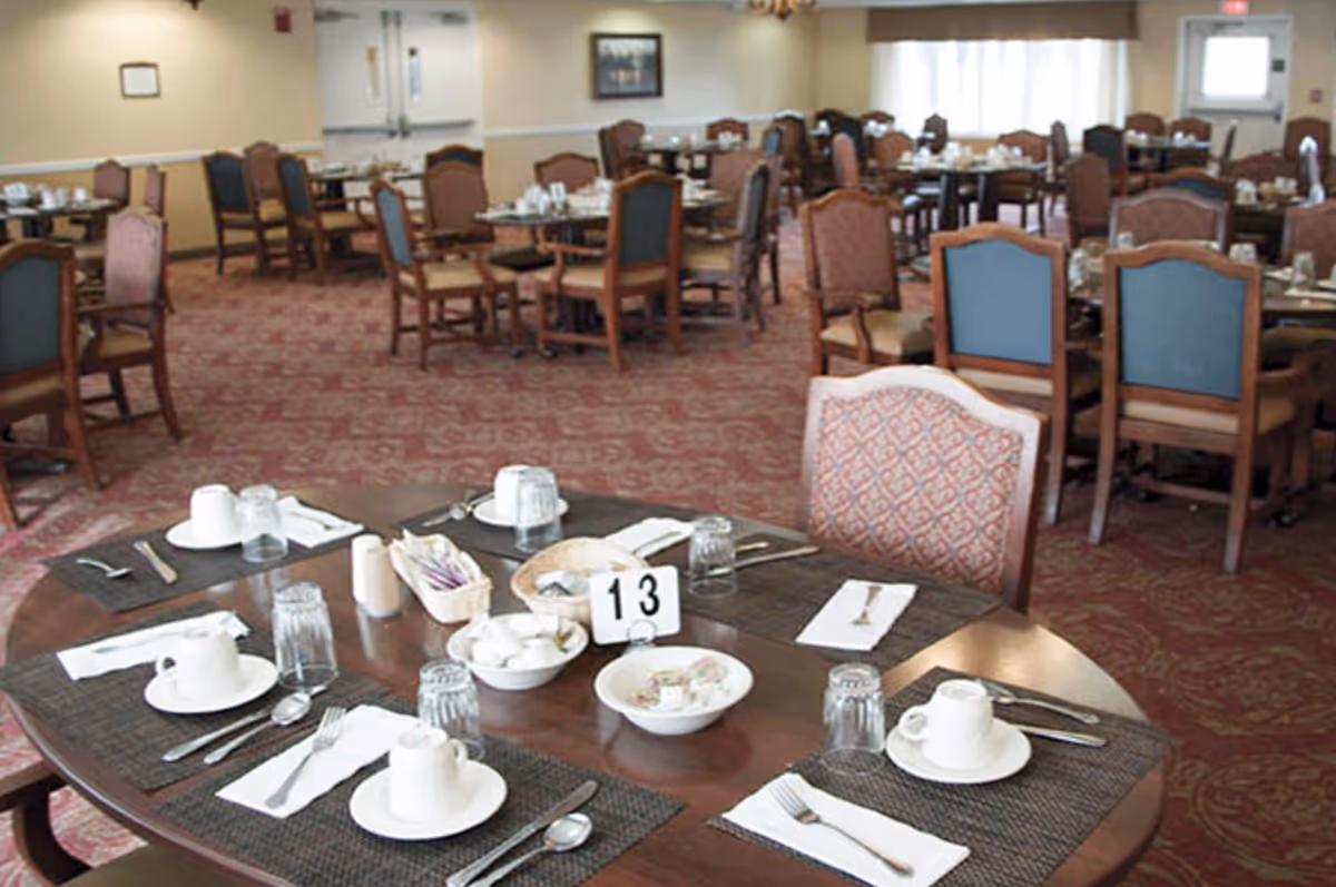A dining room in a senior living facility with multiple tables set for meals. Each table has placemats, cups, glasses, utensils, and napkins. The room has carpeted floors, upholstered chairs, and soft lighting with windows in the background.