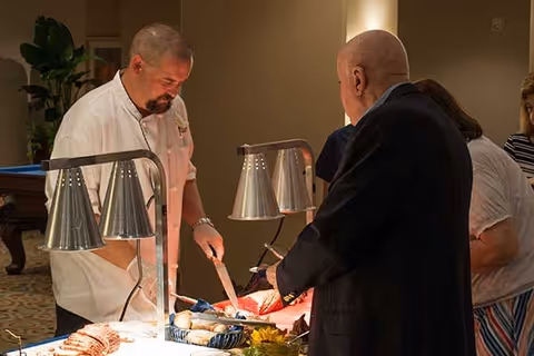 A chef serving food to an elderly man at a buffet table with heat lamps in a warmly lit room. Other people are visible in the background waiting in line.