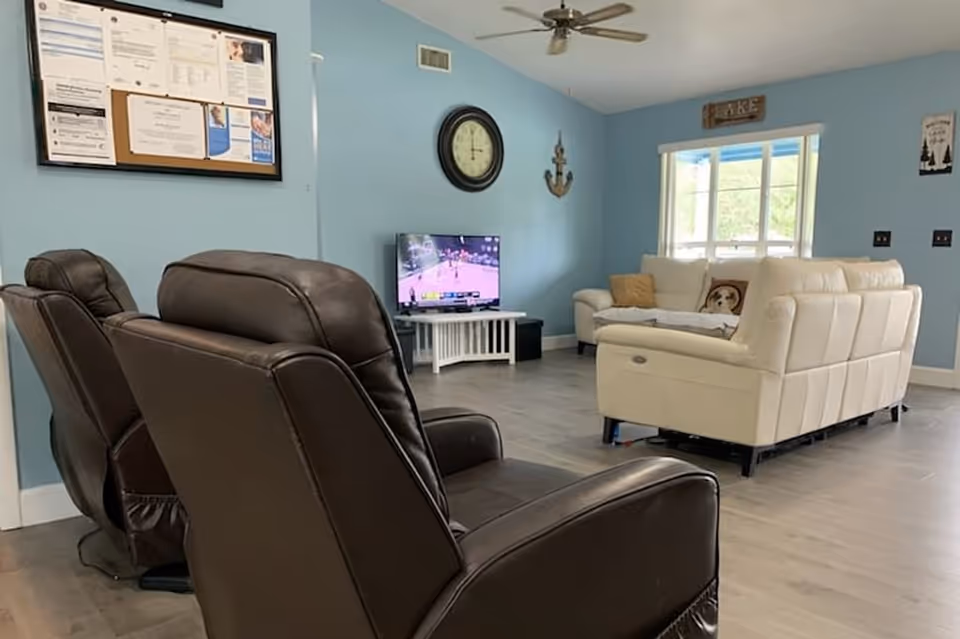A living room with light blue walls featuring two dark brown recliner chairs in the foreground and a white sectional sofa with pillows near a window. A flat-screen TV is on a white stand against the wall, showing a basketball game. A large clock and decorative anchor hang on the wall above the TV, and a bulletin board is visible on the left wall.