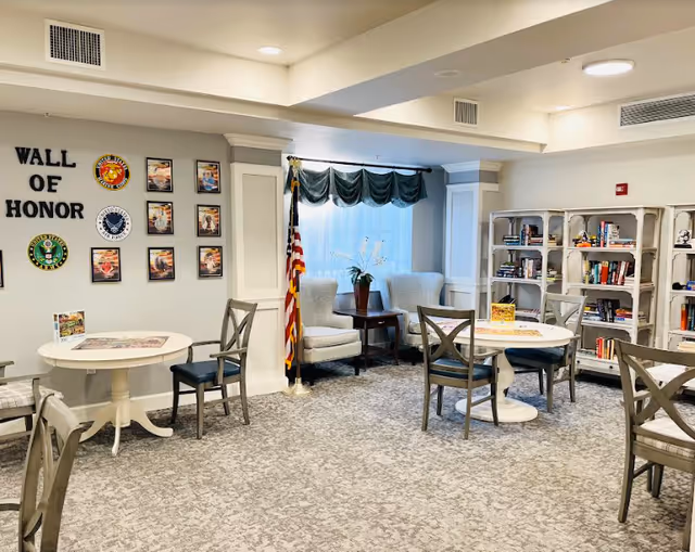 A well-lit common area with round tables and chairs, a bookshelf filled with books, two armchairs near a window with a green valance, and a Wall of Honor display featuring military emblems and photos. An American flag stands next to the armchairs.