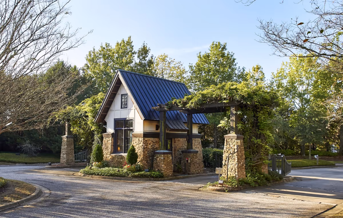 A small stone and wood guardhouse with a dark metal roof at the entrance of a facility, surrounded by greenery and trees under a clear sky.