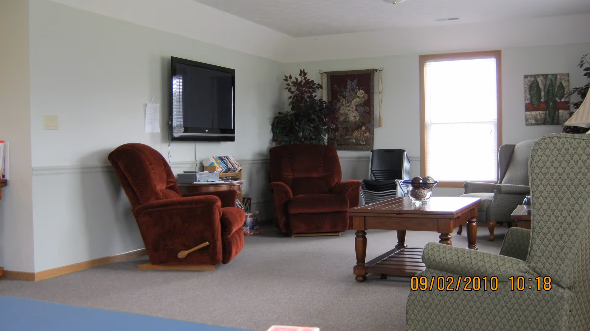 A cozy living room with two red recliner chairs, a wooden coffee table with a decorative bowl, a wall-mounted flat screen TV, a window with blinds, and additional seating including a patterned armchair and a wingback chair. The walls are light-colored with some framed artwork and a tapestry, and there are some stacked chairs in the corner.