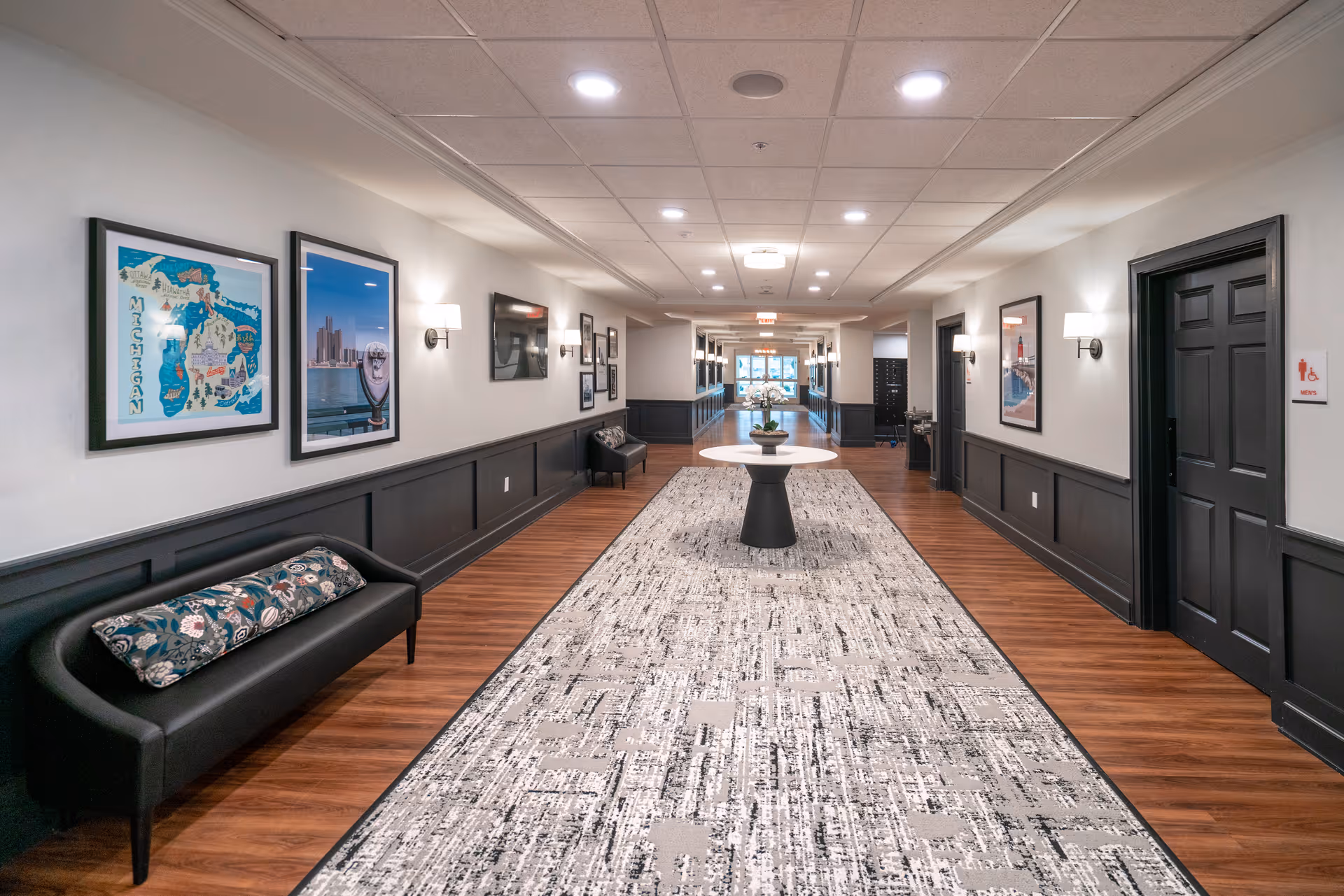 A long, well-lit hallway with wood flooring and a patterned carpet runner. The walls are painted white with dark wainscoting. There are framed pictures and wall sconces along the walls. A black bench with a decorative pillow is on the left side, and a round table with a plant is centered in the hallway. Doors and a restroom sign are visible on the right side.
