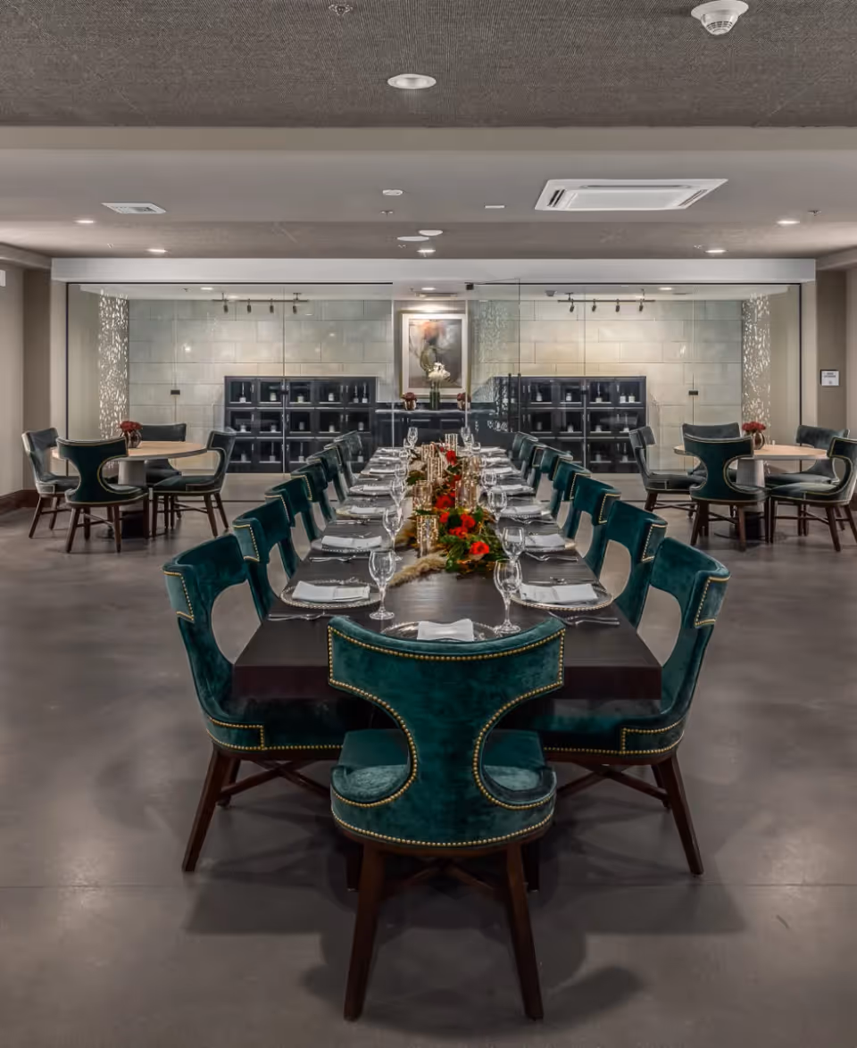 A modern dining room with a long rectangular table set for a meal. The table is surrounded by green velvet chairs with brass nailhead trim. The table is decorated with red flowers and gold candle holders. In the background, there are two smaller round tables with similar green chairs and a glass wall with shelves and a framed painting.