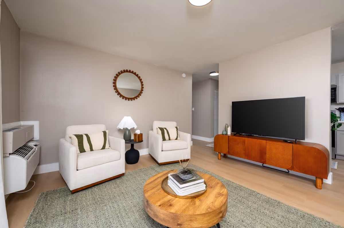 A cozy living room with two beige armchairs featuring green and white striped pillows, a round wooden coffee table with books and a decorative item on top, a round mirror with a wooden frame hanging on the wall, a small black side table with a lamp and tissue box, a wooden TV stand with a flat-screen TV, light wood flooring, and neutral-colored walls.
