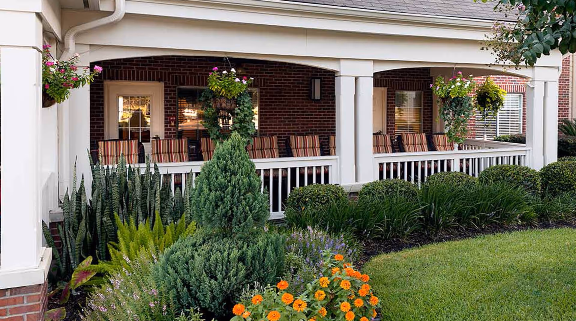 Covered brick porch with striped chairs, hanging plants, and manicured flowerbeds in front of a senior living building.