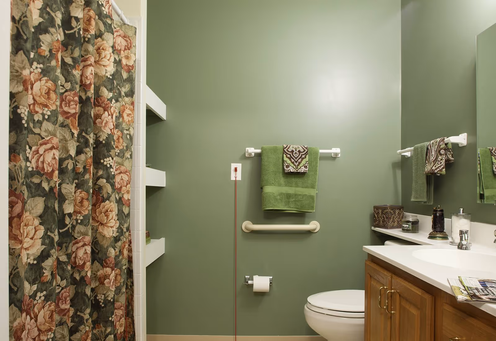 Small bathroom with green walls, a floral shower curtain, a toilet, sink vanity with mirror, and green towels.