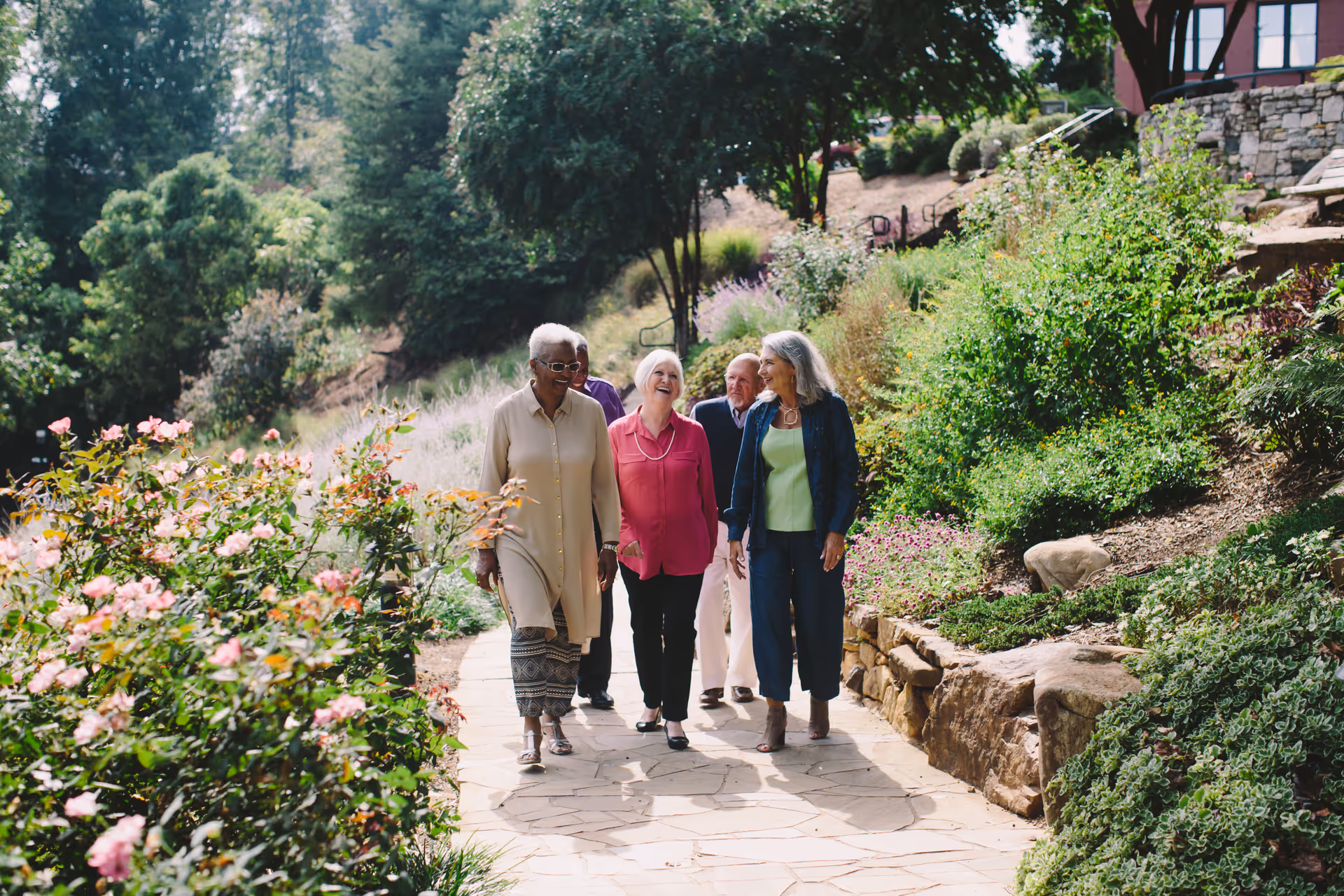 Four older adults walking and talking on a paved garden path surrounded by flowering plants and greenery.