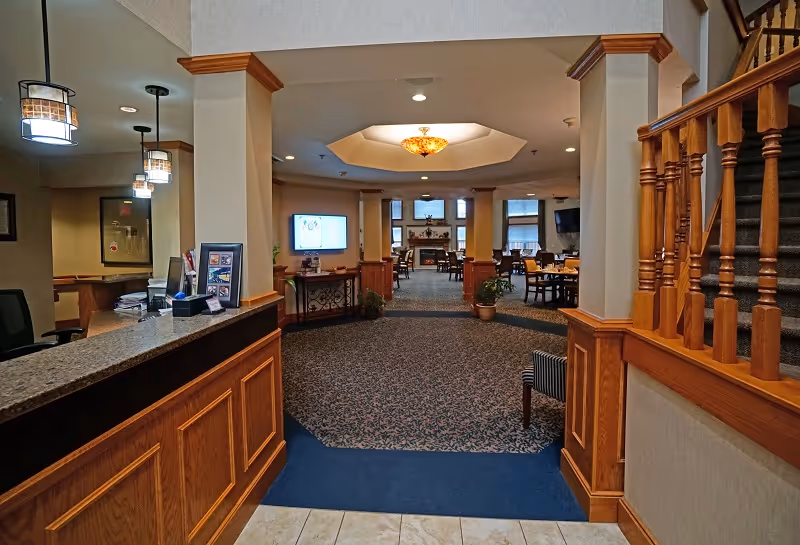 Interior view of a senior living facility lobby area with a reception desk on the left, wooden railing on the right, and a carpeted hallway leading to a dining area with tables and chairs. The ceiling features a decorative light fixture and recessed lighting.