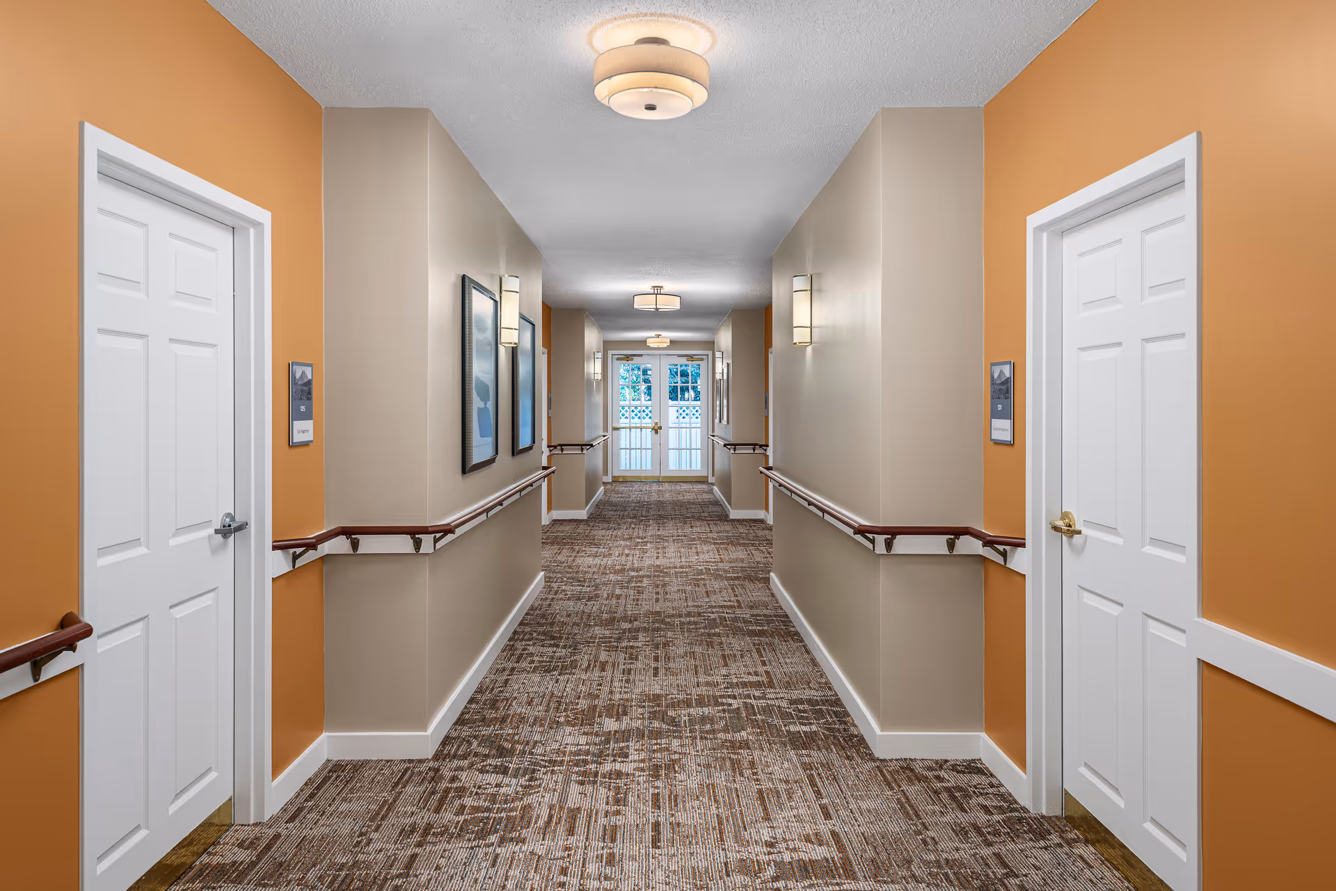 A well-lit hallway in a senior living facility with beige and orange walls, brown patterned carpet, handrails on both sides, white doors, framed artwork on the walls, and ceiling light fixtures. At the end of the hallway, there is a glass door with a view of greenery outside.
