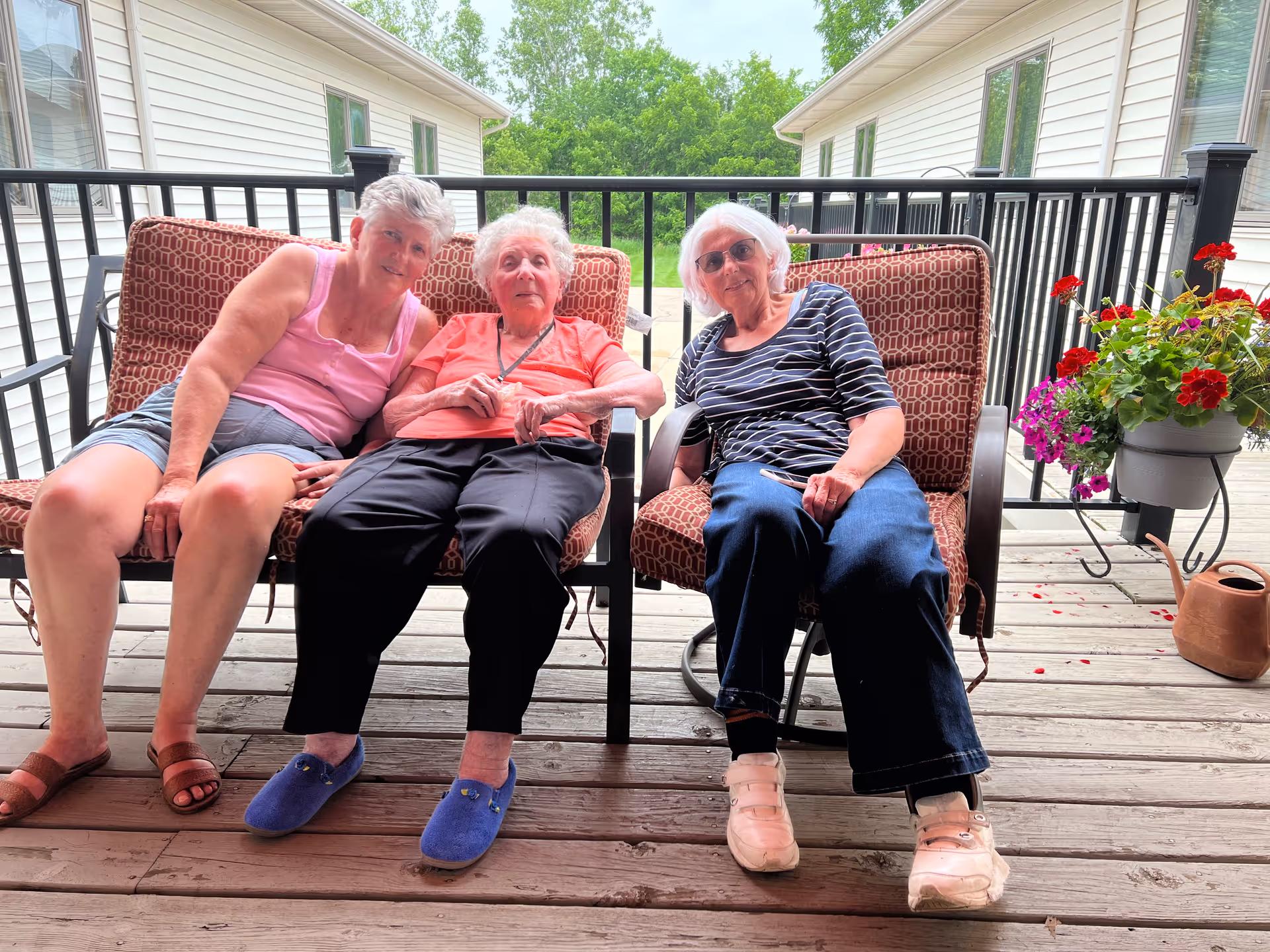 Three elderly women sitting on cushioned outdoor chairs on a wooden deck with a black railing. Behind them are two white buildings and green trees. There is a flower pot with red and purple flowers and a brown watering can on the right side of the deck.
