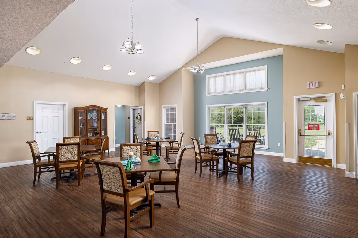 A bright dining room with several wooden tables and chairs arranged neatly. Each table has green napkins and a candle centerpiece. The room features large windows letting in natural light, light beige walls with one accent wall painted blue, and a wooden floor. There is a china cabinet against one wall and an exit door with a window on the right side.