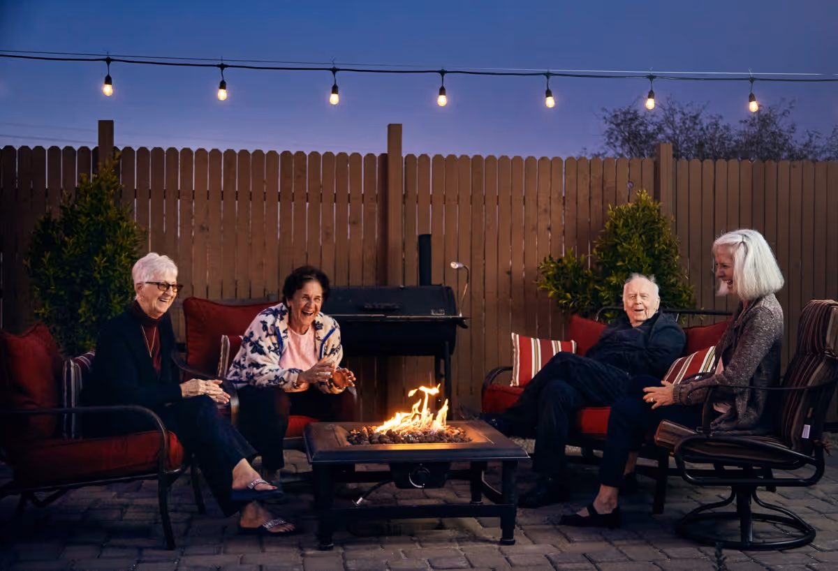 Four elderly people sitting around a fire pit in an outdoor patio area at dusk, with string lights hanging above and a wooden fence in the background. They appear to be enjoying each other's company and smiling.