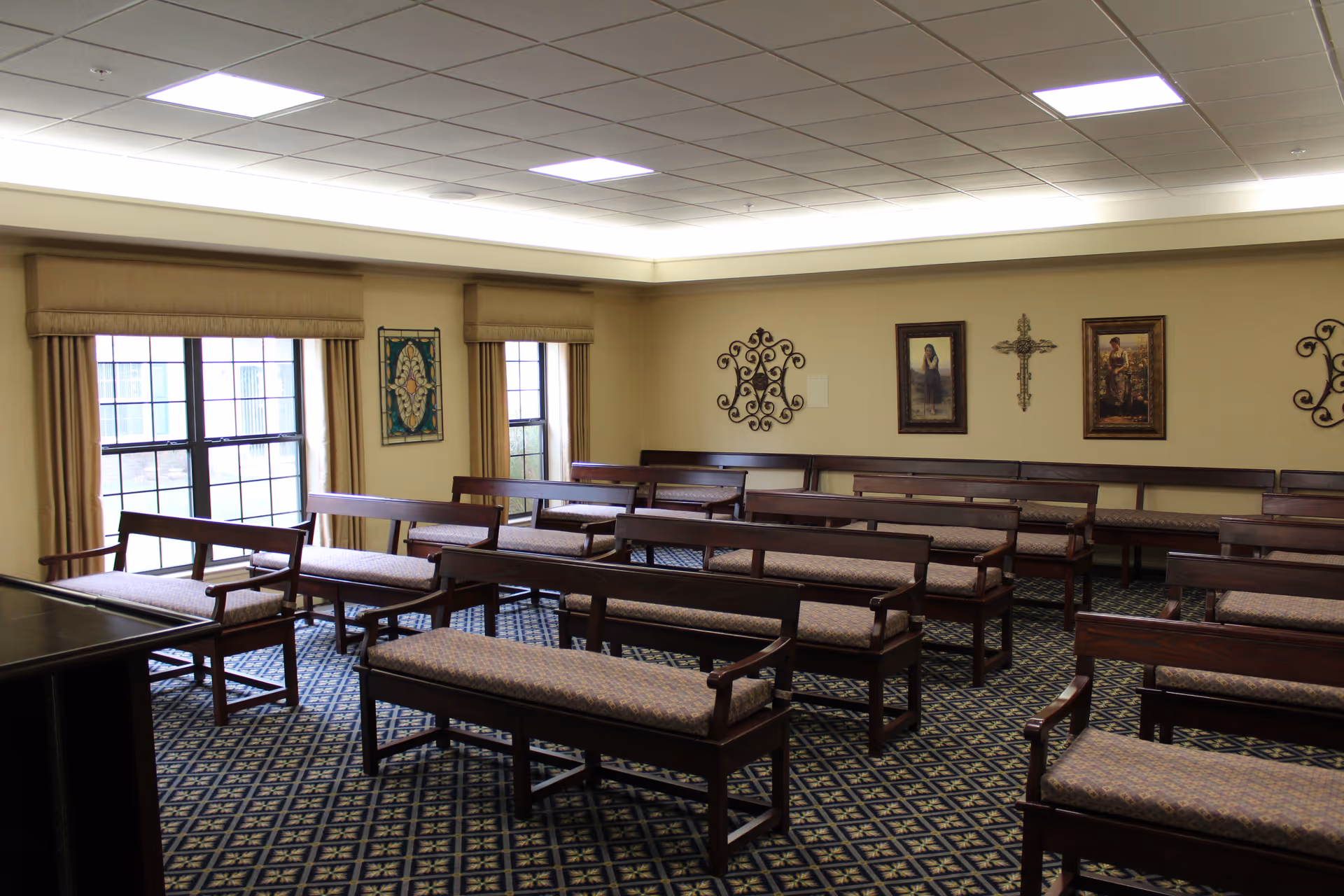 Interior view of a room with multiple wooden benches with cushioned seats arranged in rows on a patterned carpet. The room has beige walls decorated with framed artwork and a decorative cross. Large windows with beige curtains allow natural light to enter the space.