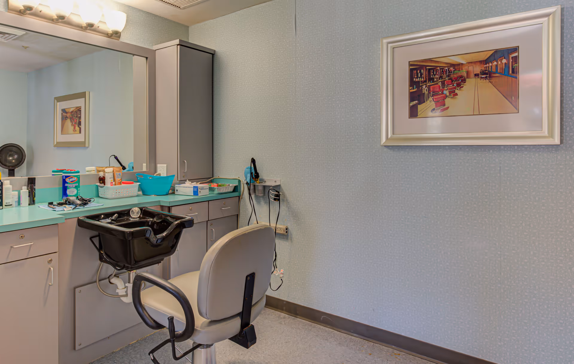 Interior view of a hair salon area in a senior living facility featuring a salon chair in front of a black hair washing sink, a large mirror, countertop with various hair care products, and a framed picture on the wall.