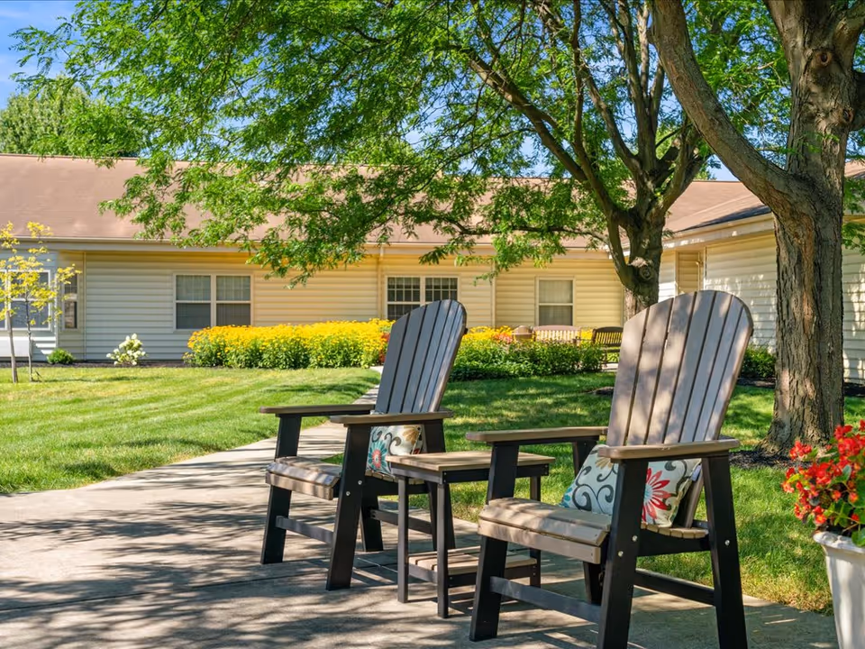Two wooden Adirondack chairs with patterned cushions sit on a paved walkway under trees in a landscaped courtyard in front of a single-story siding building.