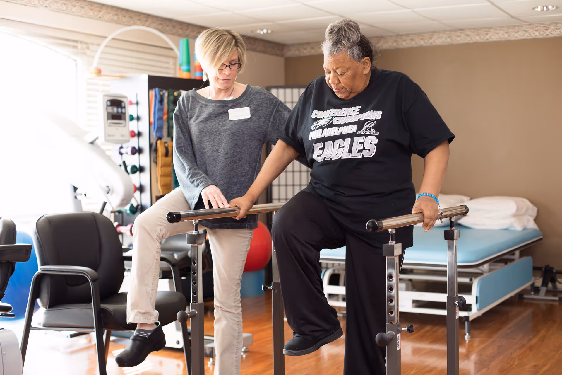 A physical therapist assists an elderly woman as she exercises by stepping over parallel bars in a therapy room with exercise equipment and a treatment table in the background.