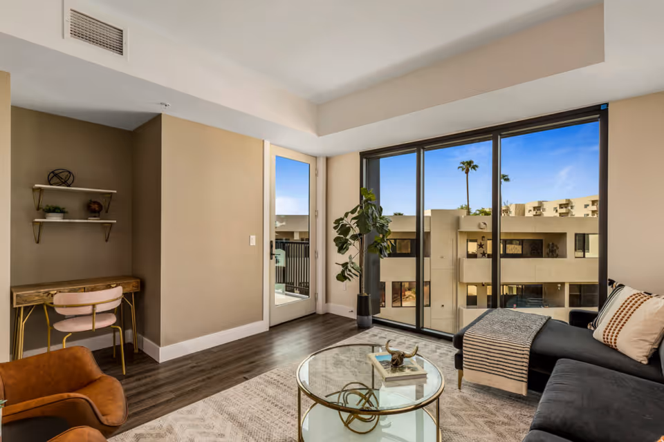 A modern living room with a large glass sliding door offering a view of an adjacent building and palm trees outside. The room features a dark sectional sofa with pillows and a throw blanket, a round glass coffee table with decorative items, a brown leather chair, a small wooden desk with a pink chair, and two wall shelves with decorative objects. The floor is dark wood, and there is a light-colored area rug.