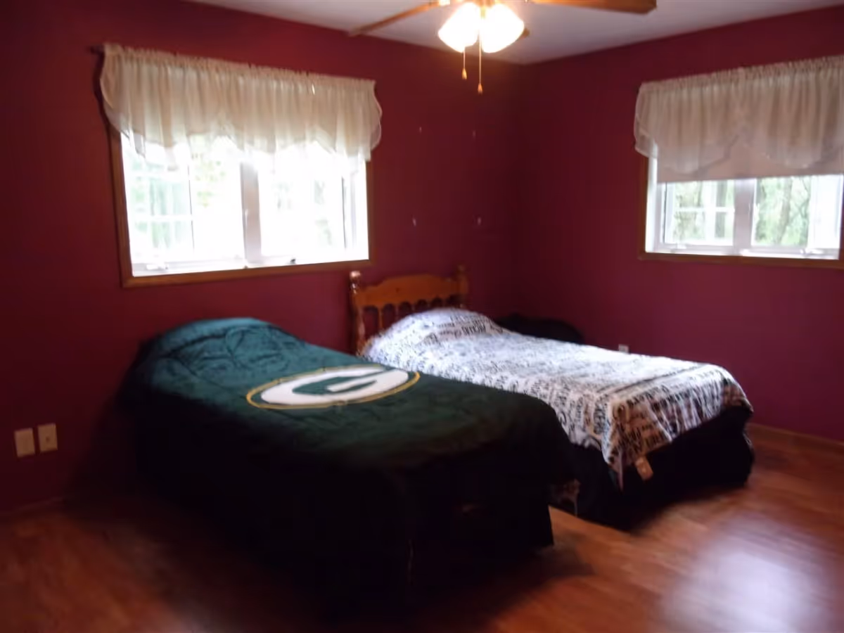 A bedroom with two single beds placed side by side against a wall with two windows. The walls are painted dark red, and the floor has wooden flooring. One bed has a green blanket with a sports logo, and the other bed has a white and black patterned blanket. There is a ceiling fan with lights above the beds.