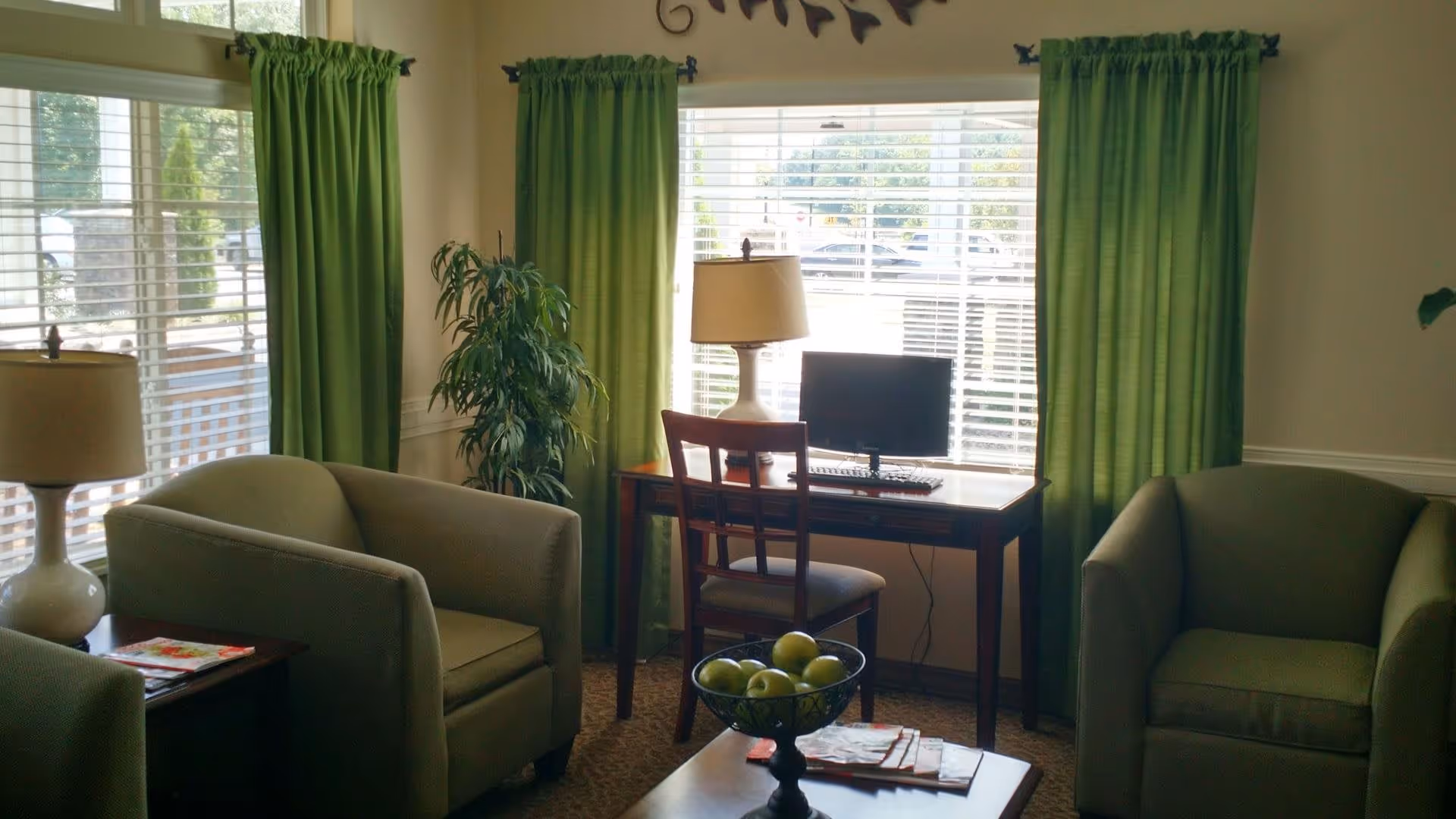 A cozy living room area with two green armchairs, a wooden desk with a chair, a computer monitor, and a lamp. The room has large windows with green curtains and a decorative plant in the corner. A bowl of green apples sits on a coffee table in the foreground.