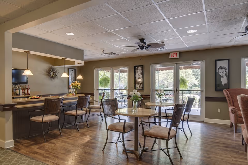 A cozy dining area in a senior living facility with round tables and chairs, a bar counter with stools, pendant lights, framed black and white portraits on the walls, ceiling fans, and large windows and doors leading to an outdoor patio with greenery visible outside.
