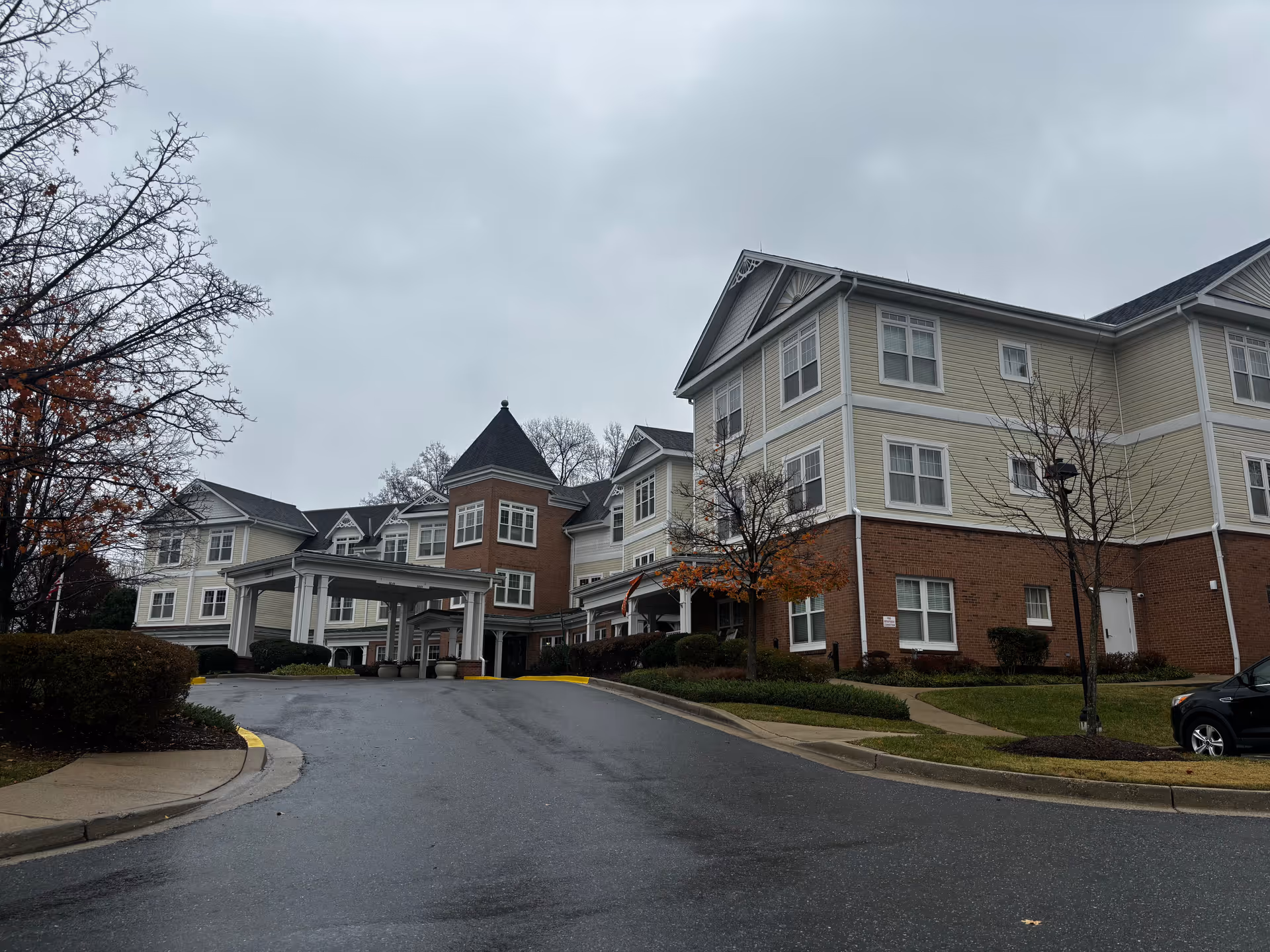 Exterior view of a multi-story senior living facility building with beige siding and red brick accents under a cloudy sky. There is a covered entrance with white pillars and a driveway leading up to it. Some trees with autumn leaves and bushes surround the building.