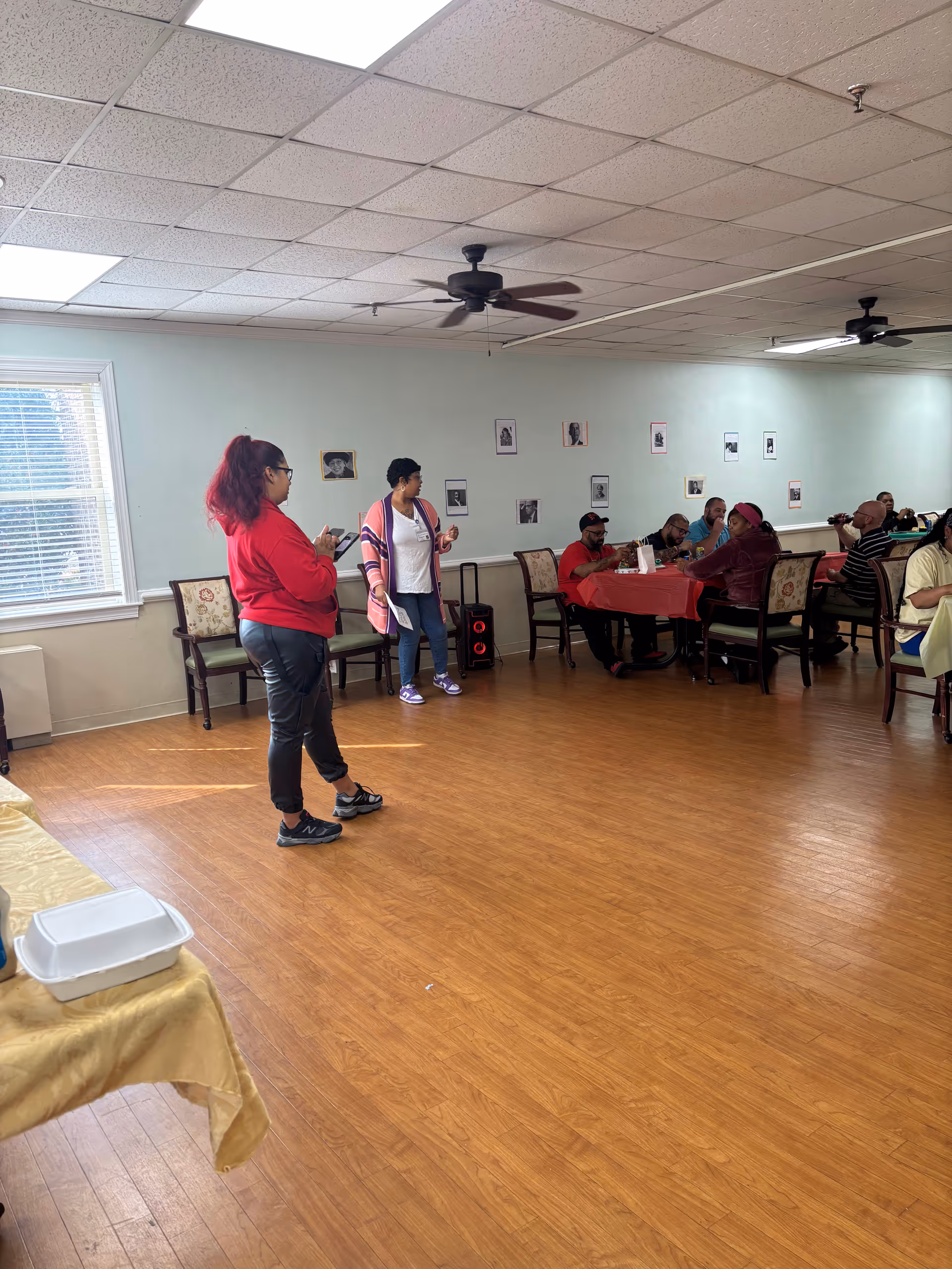 A community dining/activity room with two staff standing and several residents seated at a long table.