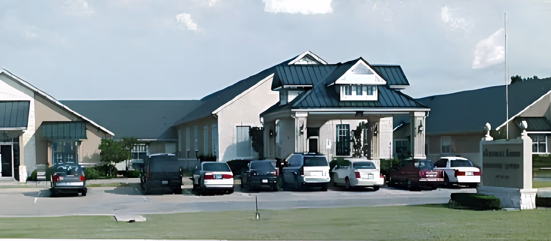 Front exterior of a single-story senior living facility with a covered entrance, parked cars, and a lawn.