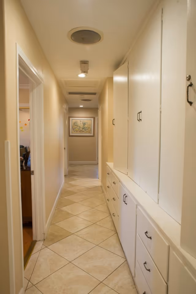 A narrow hallway with beige walls and tiled floor. On the right side, there are white built-in cabinets and drawers with black handles. At the end of the hallway, a framed picture is hanging on the wall. The left side shows an open doorway leading to another room.