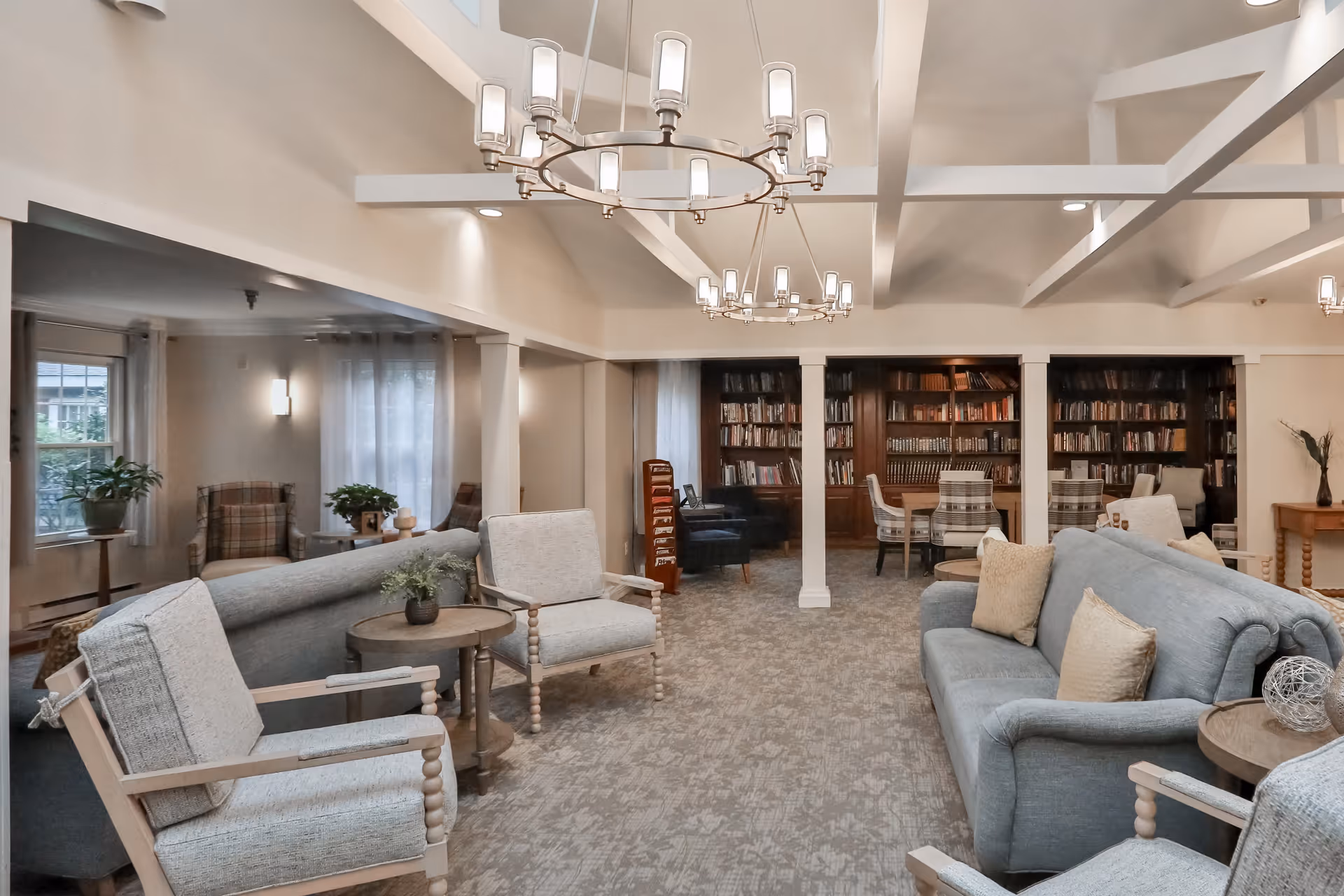 A spacious senior living common area with comfortable seating including sofas and armchairs arranged around small tables. The room features a carpeted floor, large windows with curtains, and built-in bookshelves filled with books. Modern chandeliers hang from the ceiling, providing warm lighting.