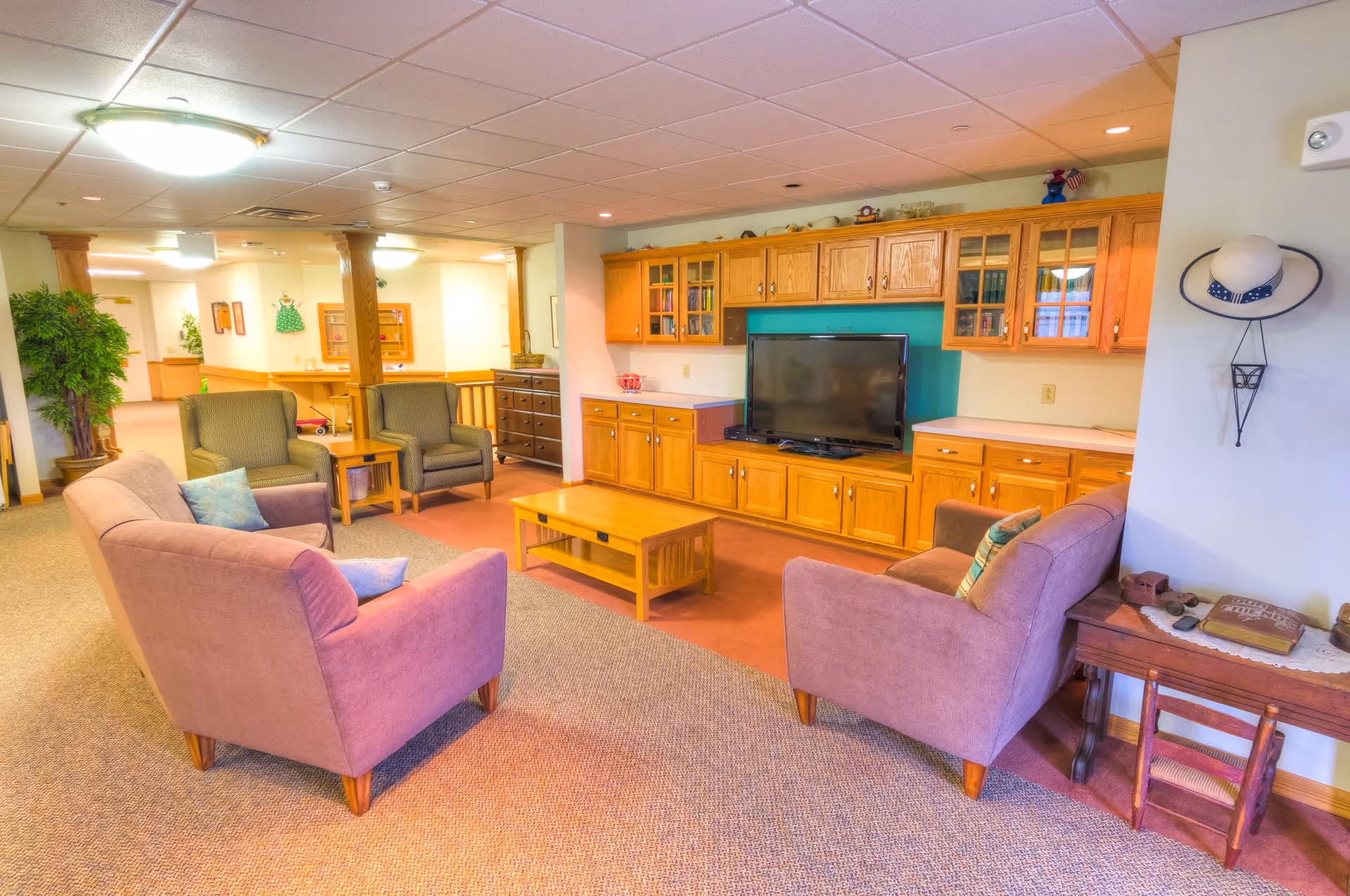 A cozy living room area in a senior living facility with two purple armchairs, two green armchairs, a wooden coffee table, and a large wooden cabinet with a flat-screen TV. The room has carpeted floors, a ceiling light, and decorative items including a hat on the wall and plants in the background.