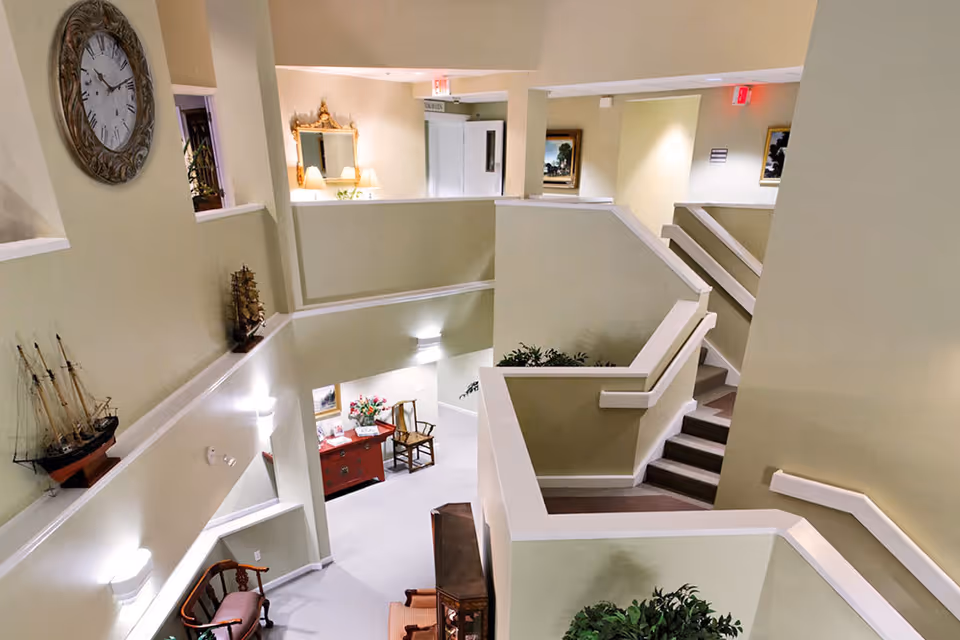Interior view of a multi-level senior living facility with beige walls, white railings, and carpeted stairs. The area features decorative elements including a large wall clock, model ships, framed paintings, a mirror with lamps, and various chairs and tables. The space is well-lit with wall sconces and ceiling lights.