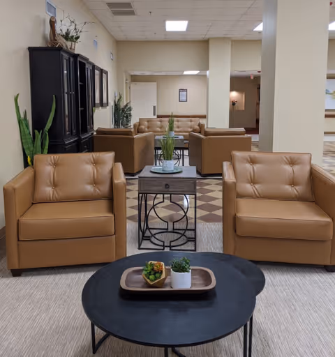 A seating area in a nursing home featuring two tan leather armchairs in the foreground, a black round coffee table with small decorative plants, and additional tan leather sofas and armchairs arranged around a side table with a plant. The room has beige walls, a checkered floor pattern in the background, and ceiling lights.