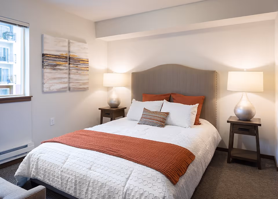 A neatly made bed with a beige upholstered headboard, white textured bedding, and rust-colored pillows and throw blanket. Two wooden nightstands with silver lamps flank the bed. A window on the left side shows part of an adjacent building, and abstract artwork hangs on the wall above the left nightstand.