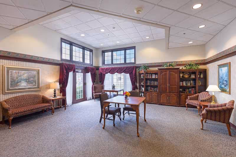 A spacious living room area with carpeted floor, featuring a wooden table with four chairs in the center. The room has large windows with burgundy curtains and valances, allowing natural light to enter. There are two sofas and two armchairs upholstered in patterned fabric, a wooden bookshelf filled with books and decorative plants on top, and framed paintings on the walls. Two table lamps provide additional lighting.