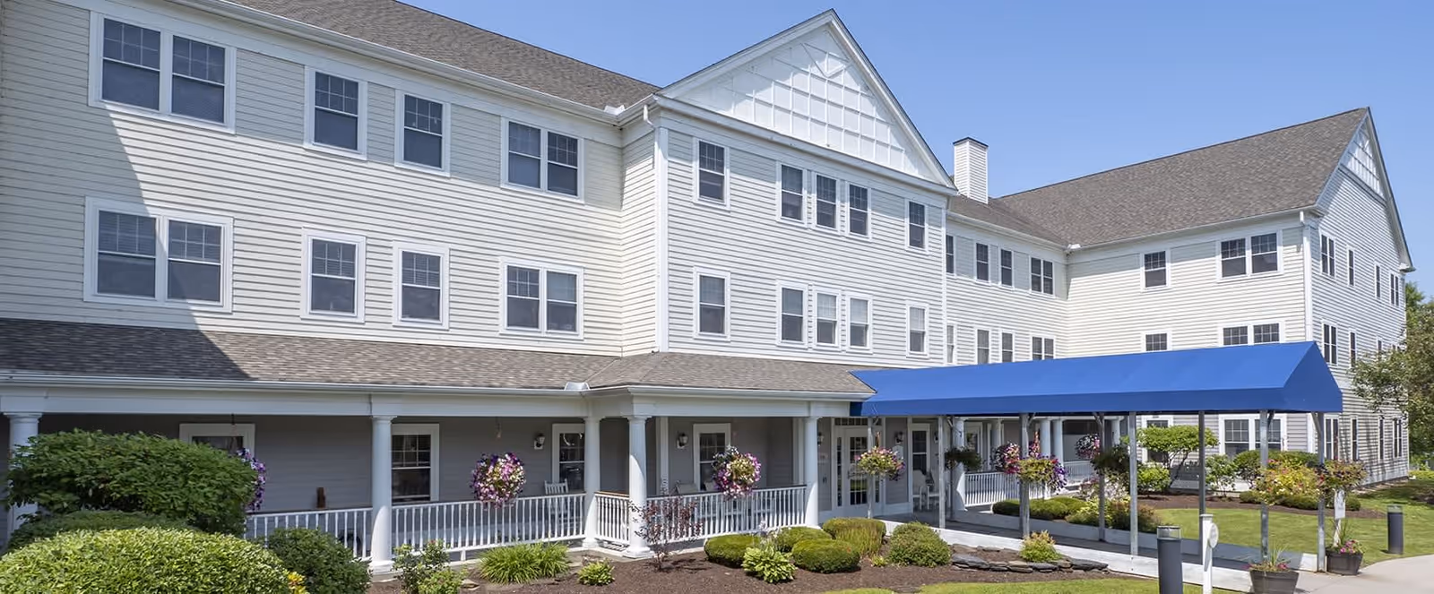 Exterior view of a large, three-story senior living facility building with white siding and multiple windows. The entrance features a covered walkway with a blue canopy and hanging flower baskets. Well-maintained landscaping with bushes and flowers surrounds the building under a clear blue sky.