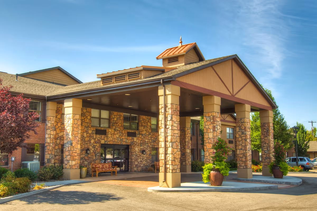 Exterior view of a senior living facility entrance with a covered drop-off area supported by stone pillars. There are benches and large potted plants near the entrance, and the building has a combination of stone and brick facade under a clear blue sky.