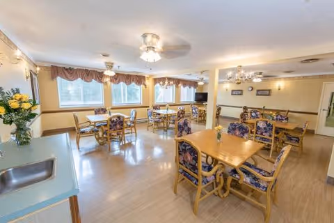 Bright dining room with several wooden tables and floral-upholstered chairs, large windows with valances, ceiling fans, and floral arrangements.