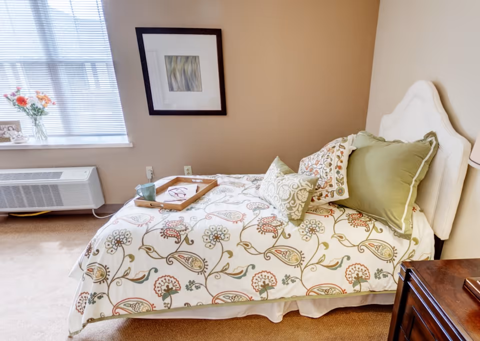 A cozy bedroom with a single bed featuring a floral patterned bedspread and multiple pillows. A wooden tray with a green mug and a pair of glasses rests on the bed. There is a window with blinds partially open, a vase with flowers on the windowsill, a framed abstract artwork on the wall, and a wooden dresser partially visible on the right.