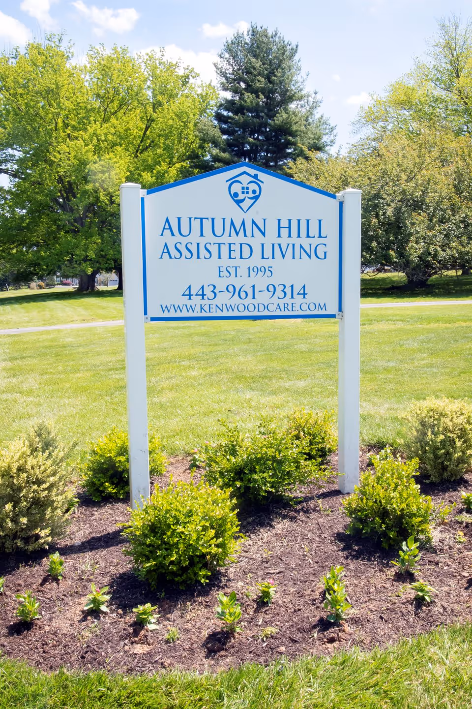 A white and blue sign for Autumn Hill Assisted Living with contact information, surrounded by small green bushes and grass, with trees and a clear sky in the background.